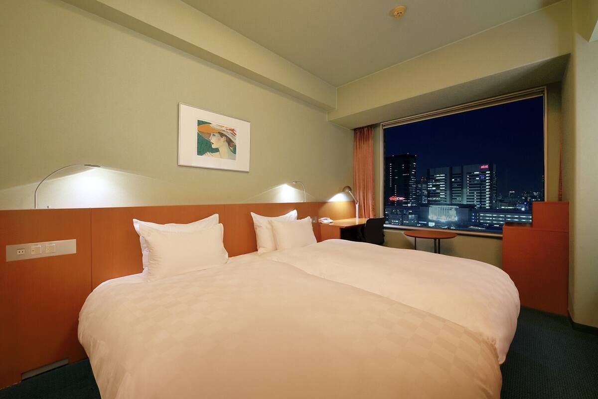 A hotel room with a large bed, two bedside lamps, a night city view, and a framed artwork on the wall.