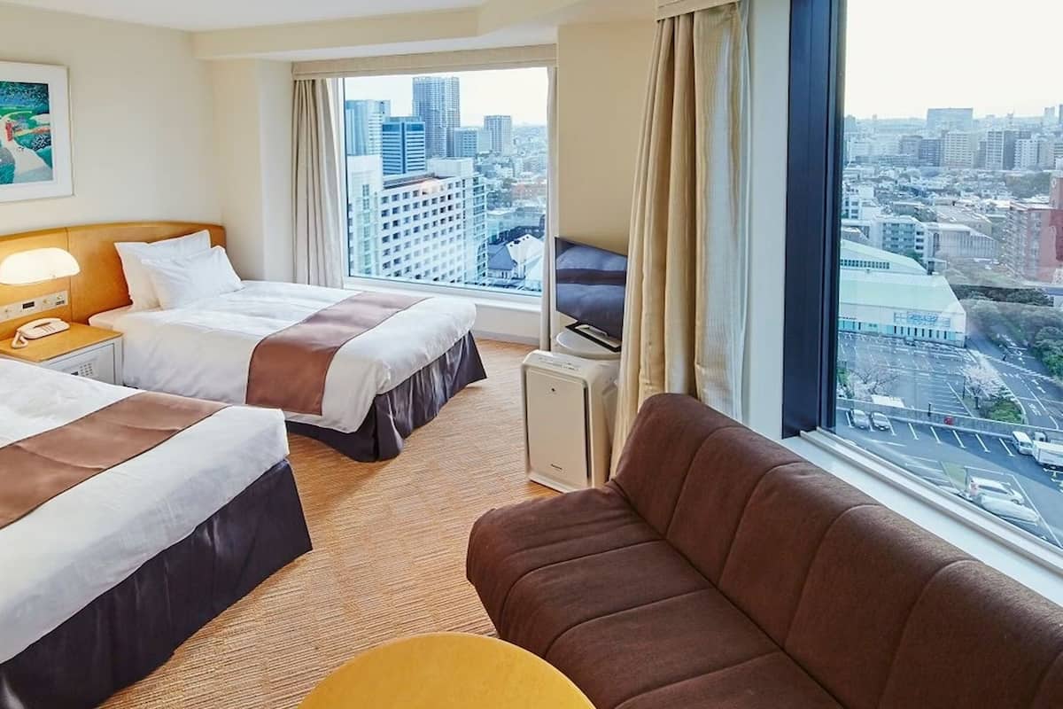 A hotel room with two beds, a sofa, a small table, and a large window with a city view.