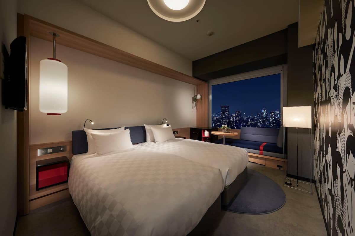 A hotel room with a large bed, bedside tables, a TV, a window with a city view, and a wall with a comic strip design.