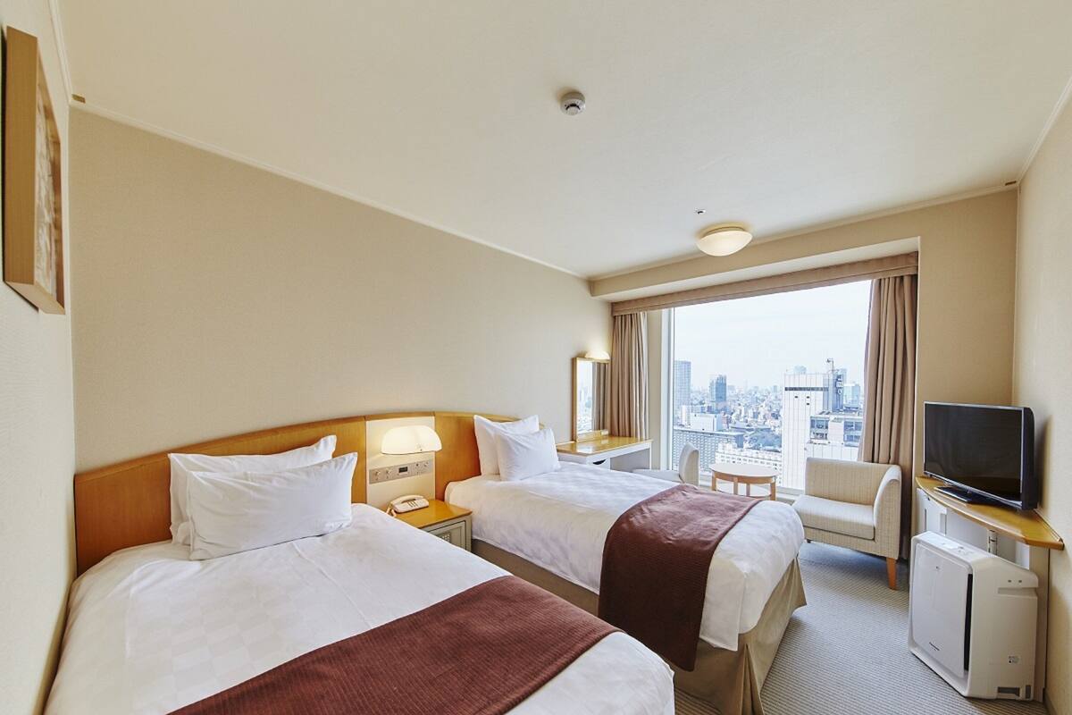 A hotel room with two beds, a desk, a chair, a television, and a view of the city.