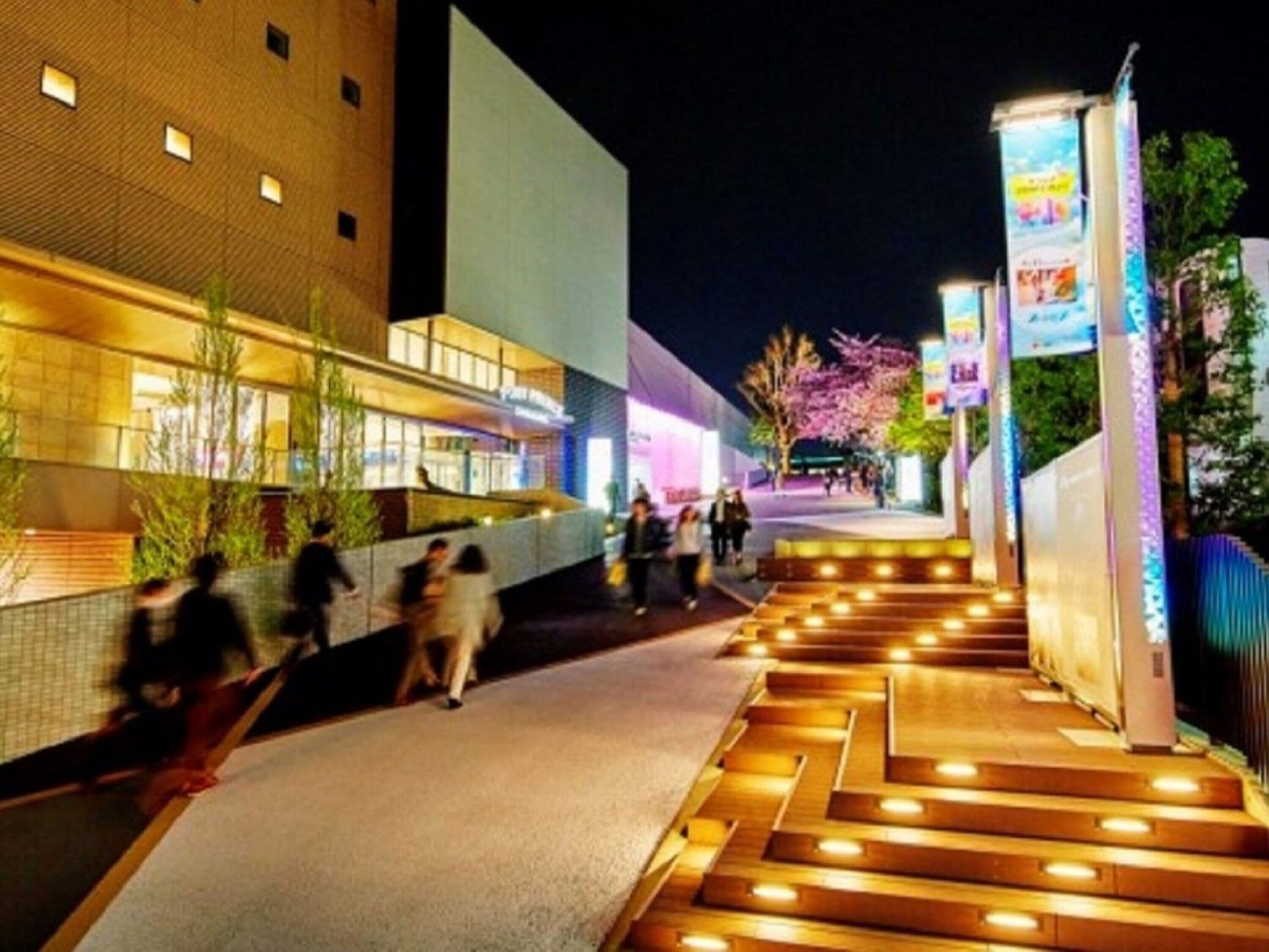 A nighttime urban scene with a modern building, a pedestrian walkway, and illuminated signage.
