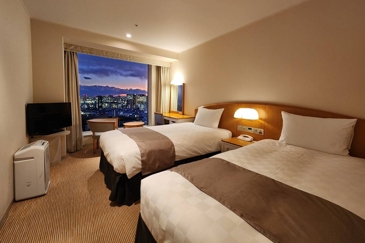 A hotel room with two beds, a desk, a TV, and a view of a cityscape at dusk.