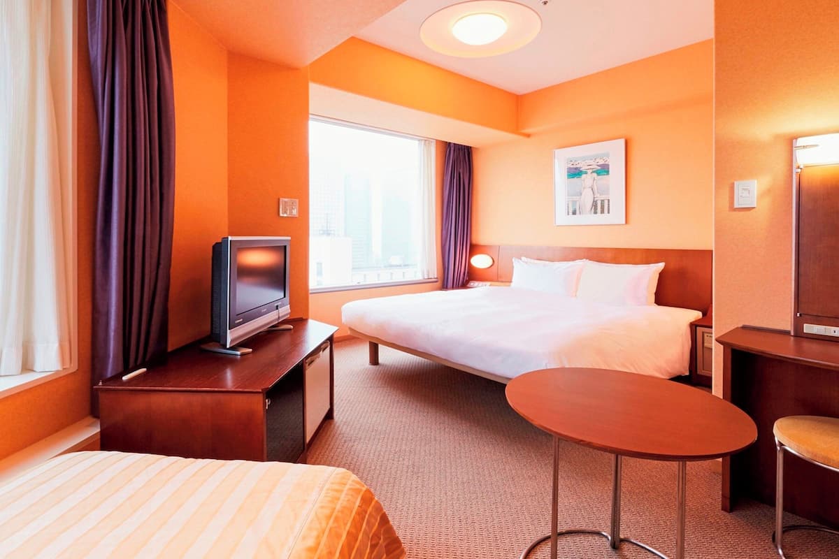A hotel room with a bed, a desk with a television, a small table, and a window with curtains.