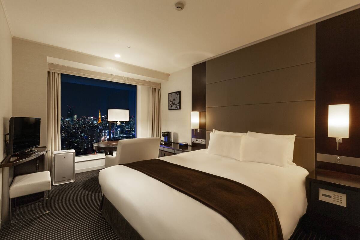 A hotel room with a large bed, a desk with a computer, a chair, a TV, and a view of the city at night.