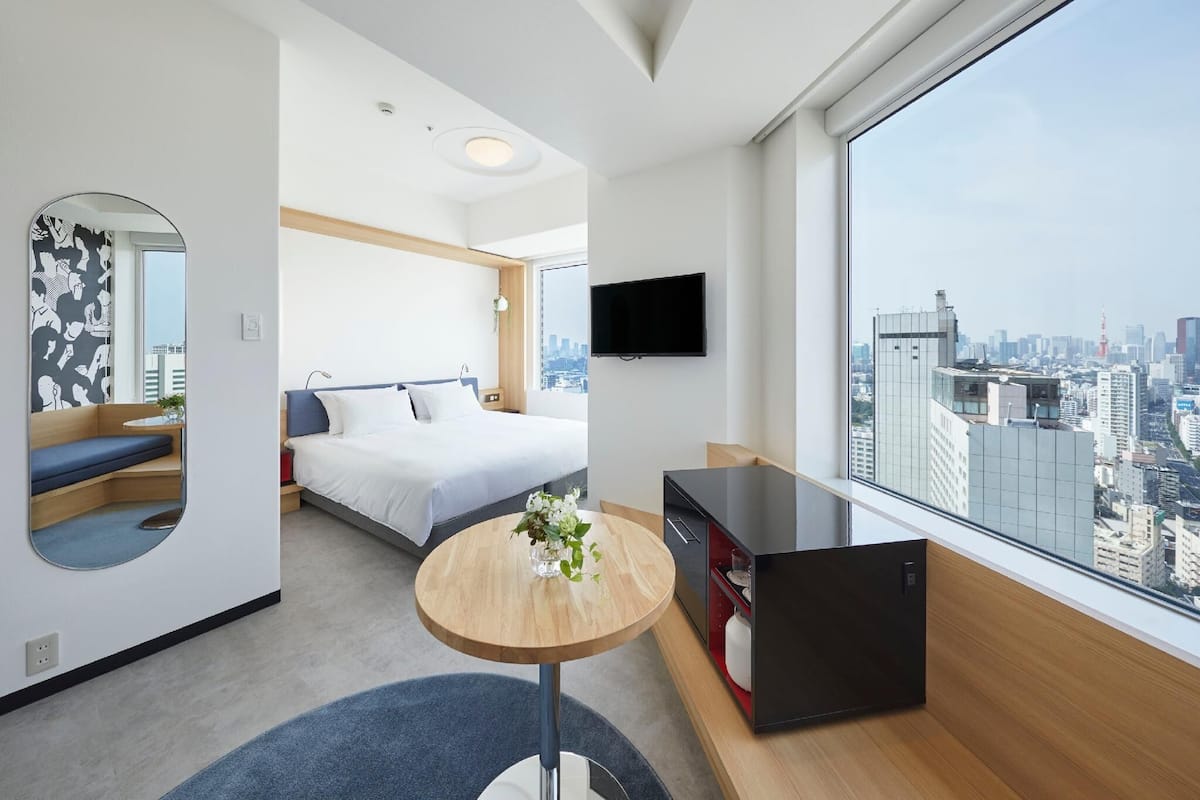 A modern hotel room with a bed, a round table with a vase, a TV, and a city view.