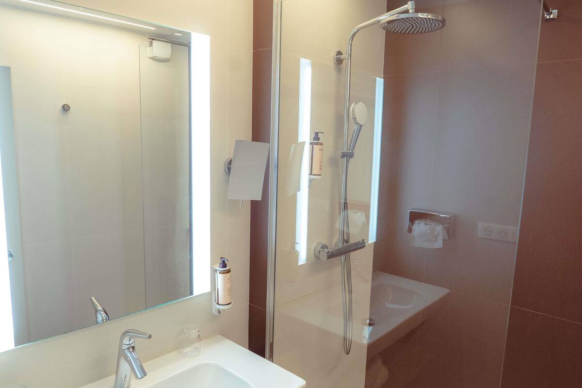 Un baño moderno con un espejo grande, un lavamanos, una ducha con cabezal de ducha manual y zona de ducha con paredes de vidrio.