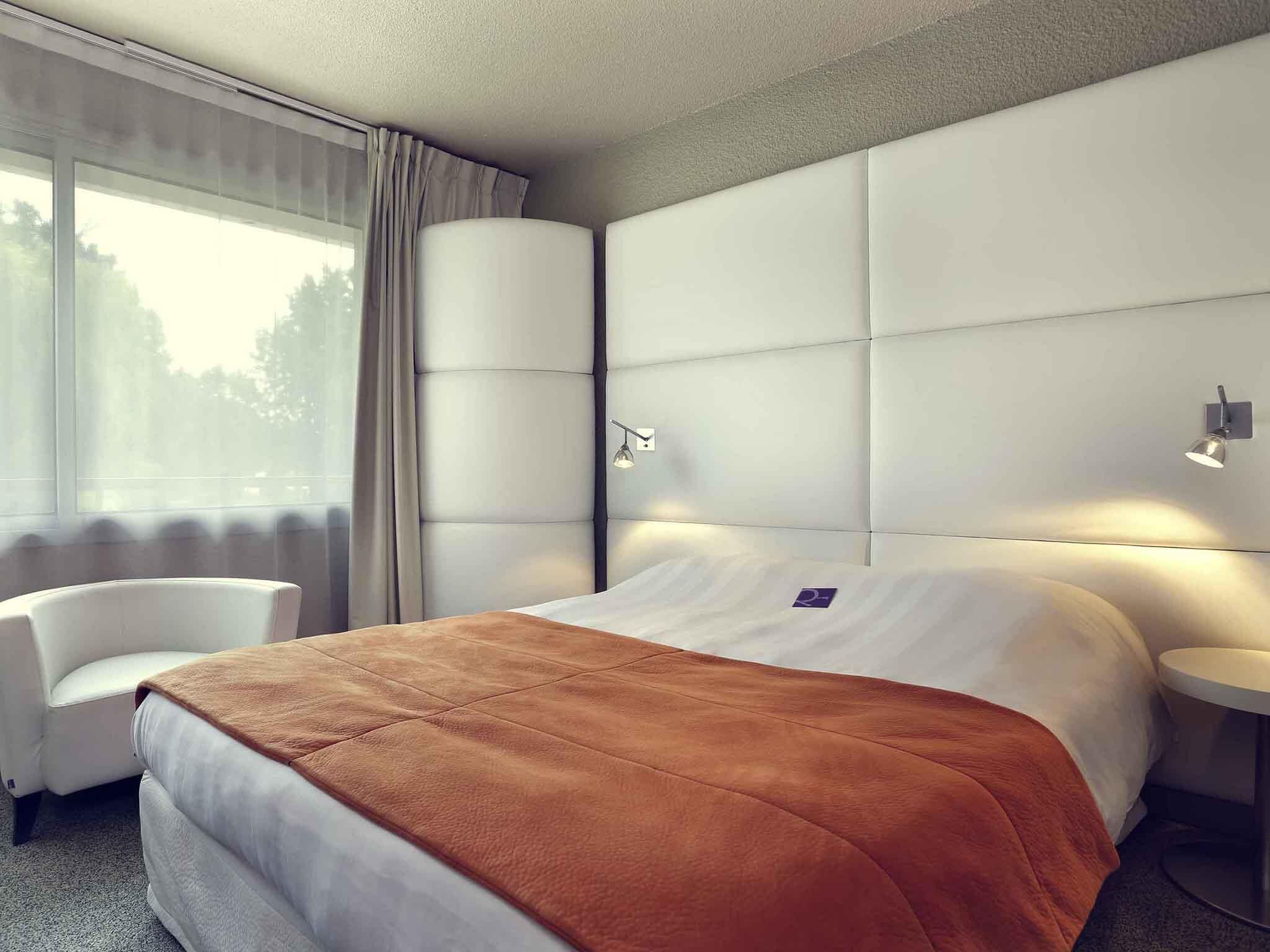 A modern hotel room with a large bed, a chair, a bedside table, and a window with curtains.