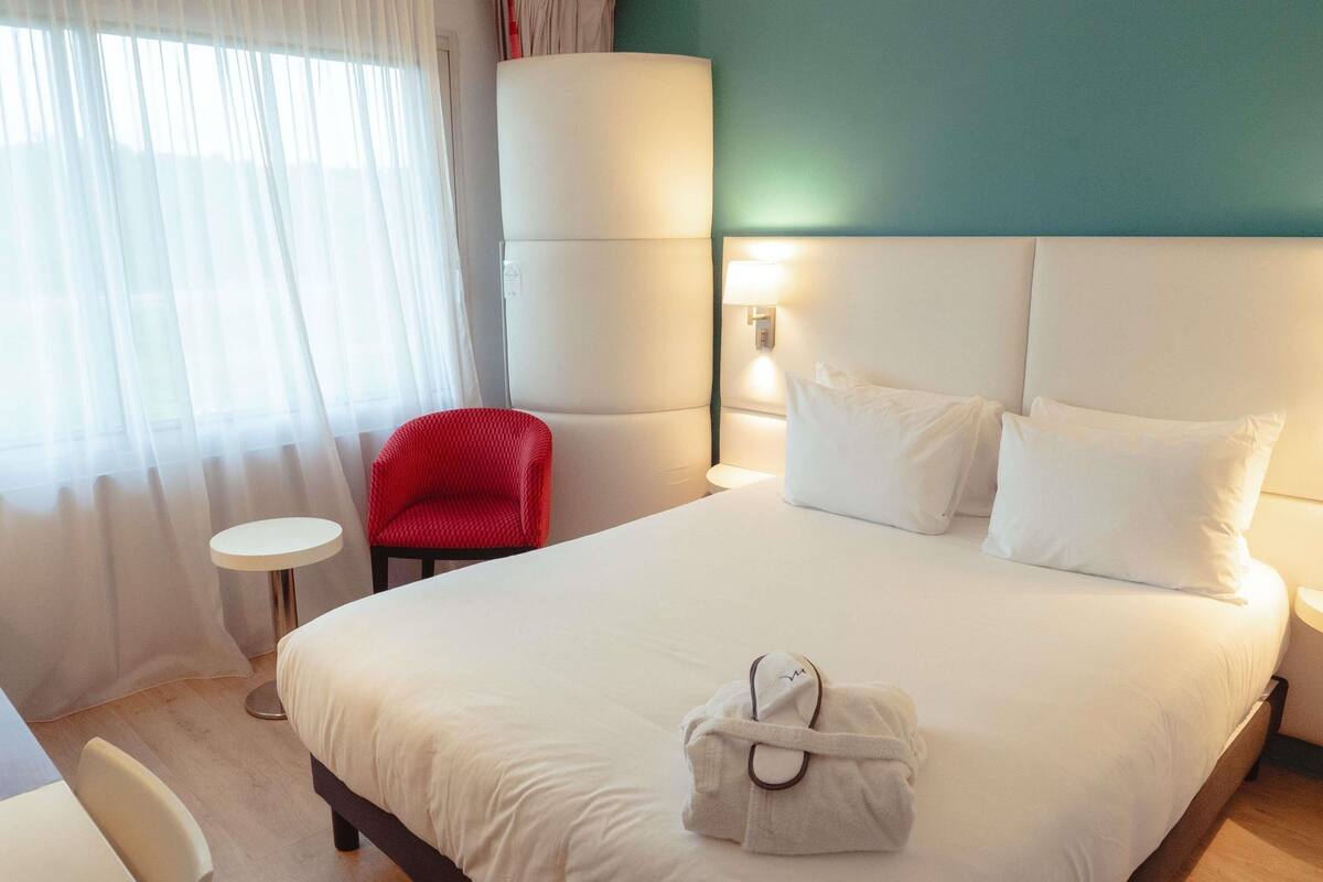 A hotel room with a bed, a red chair, a small round table, and a wall-mounted lamp.