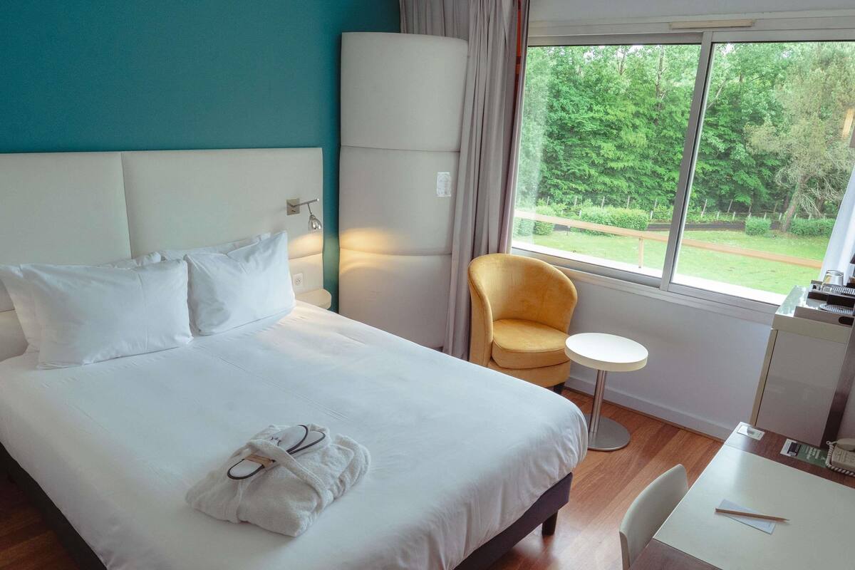 Habitación de hotel con cama, una silla, una mesita y ventana con vistas a zonas verdes.