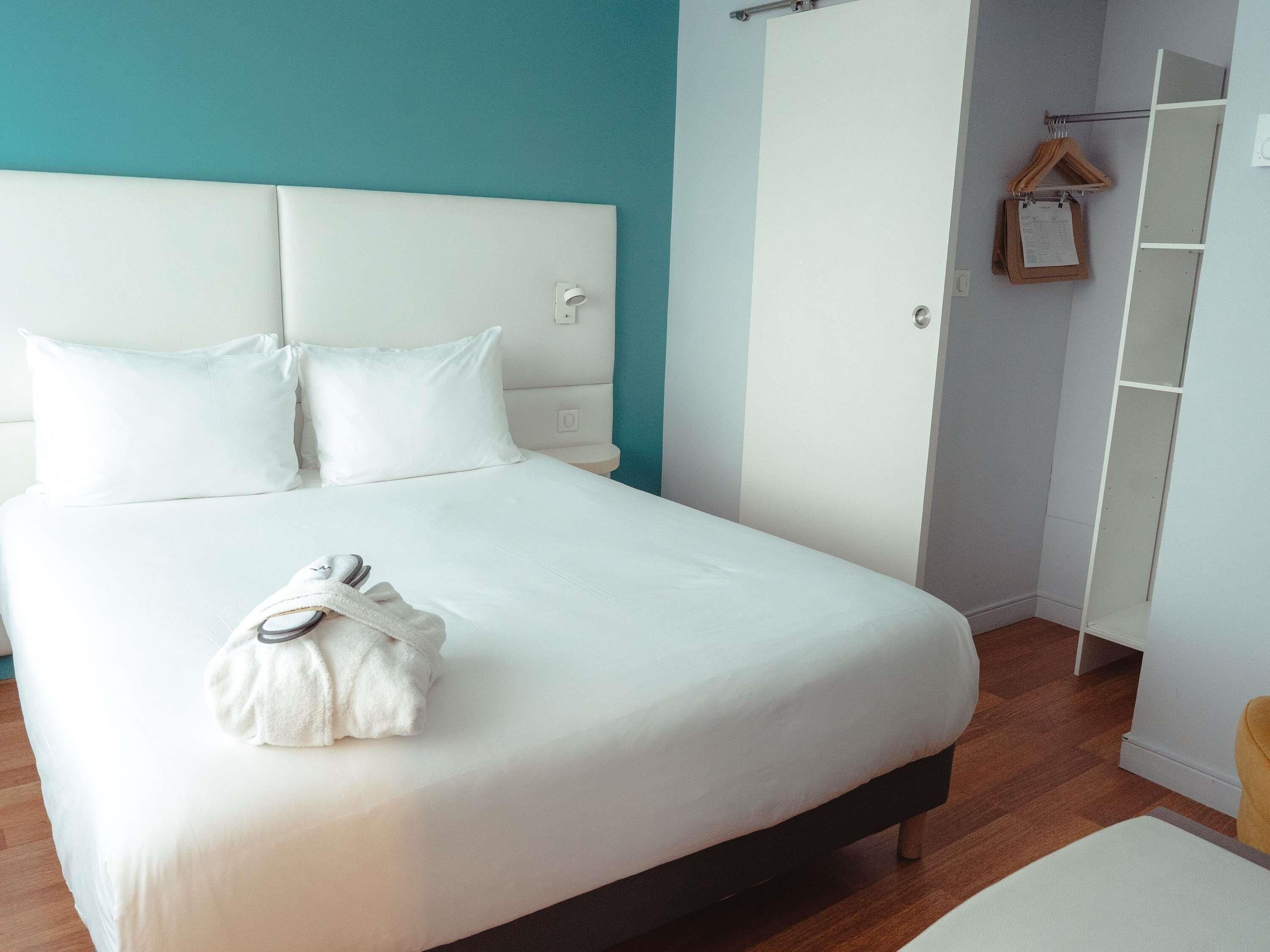 Un lit bien fait, avec du linge de lit blanc et une tête de lit blanche, un plancher en bois et un mur d’accent de couleur turquoise.
