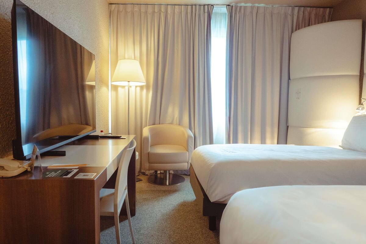 Una habitación de hotel con escritorio, silla, dos camas, televisor y cortinas.
