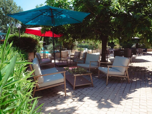 An outdoor seating area with chairs and tables, a pool in the background, and umbrellas providing shade.