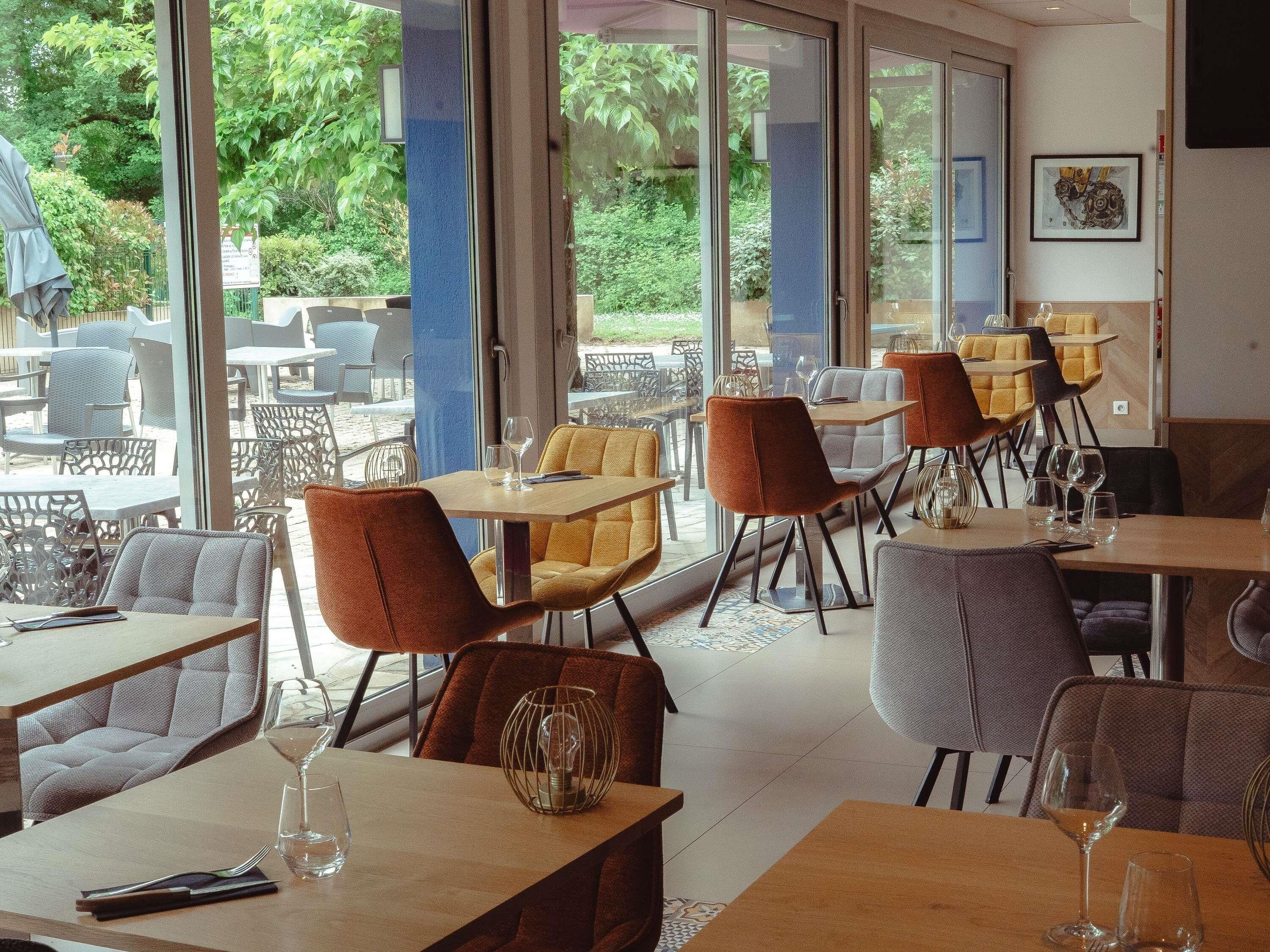 An indoor dining area with tables and chairs, large windows, and a view of greenery outside.