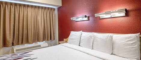 A hotel room with a bed, curtains, a wall-mounted air conditioning unit, and a light fixture.
