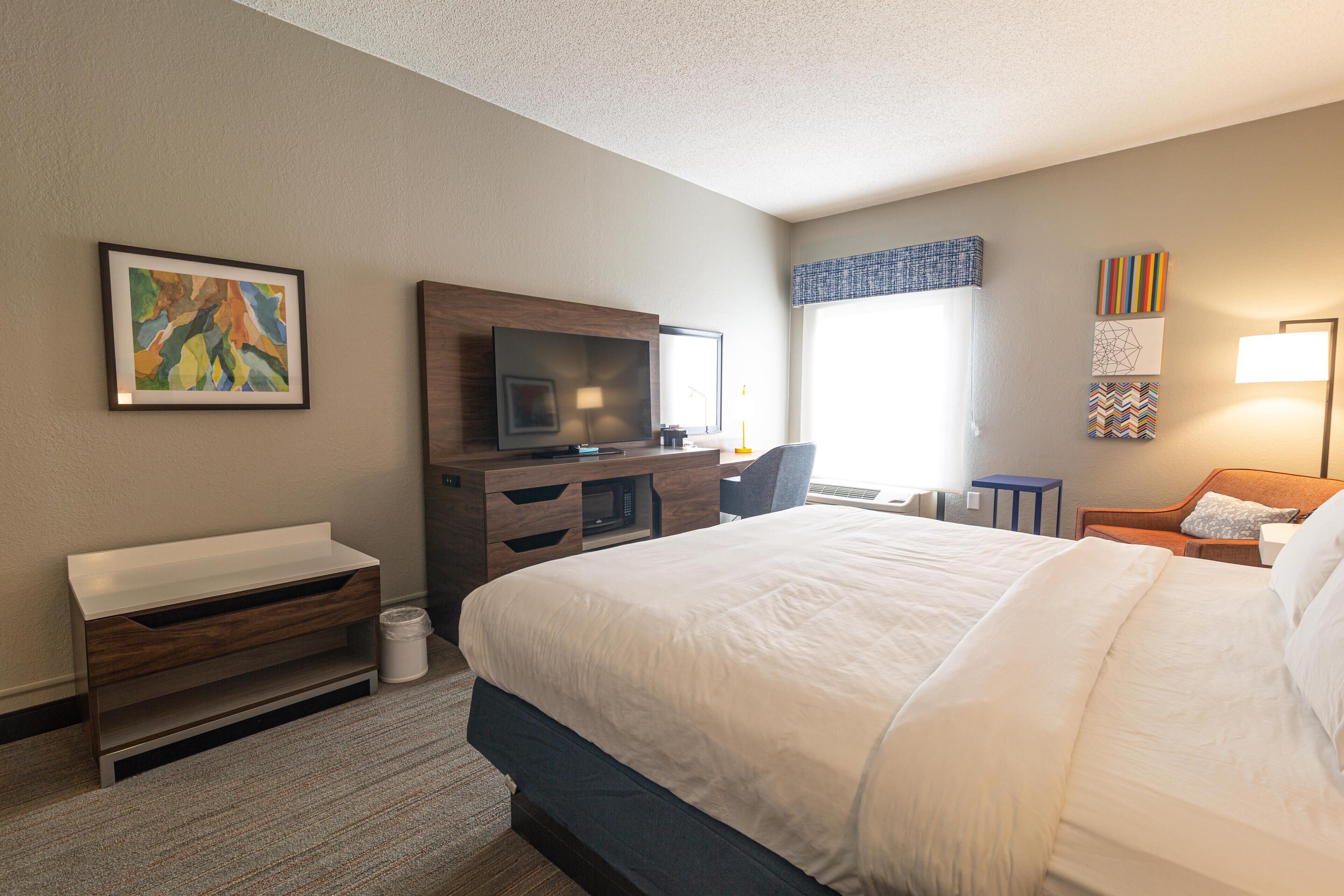 A hotel room with a bed, a desk with a TV, a small table, and artwork on the walls.