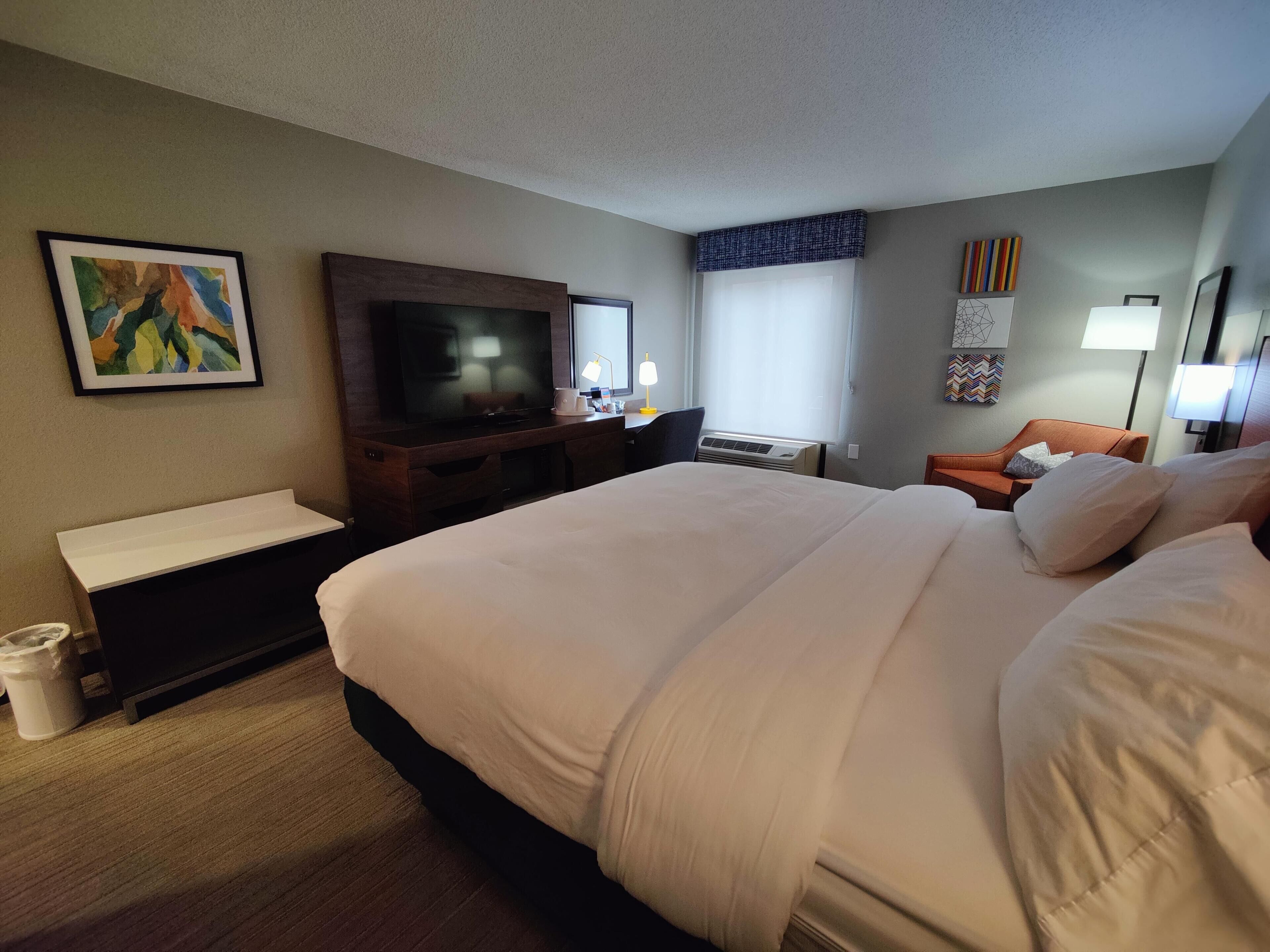 A hotel room with a large bed, a flat-screen TV, a desk with a lamp, and a window with blinds.
