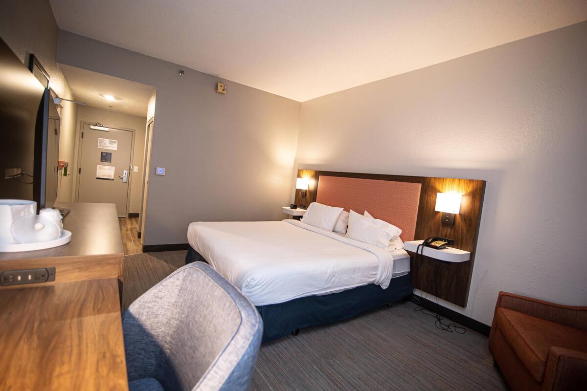 A hotel room with a bed, bedside table, chair, and a desk with a cup and saucer.