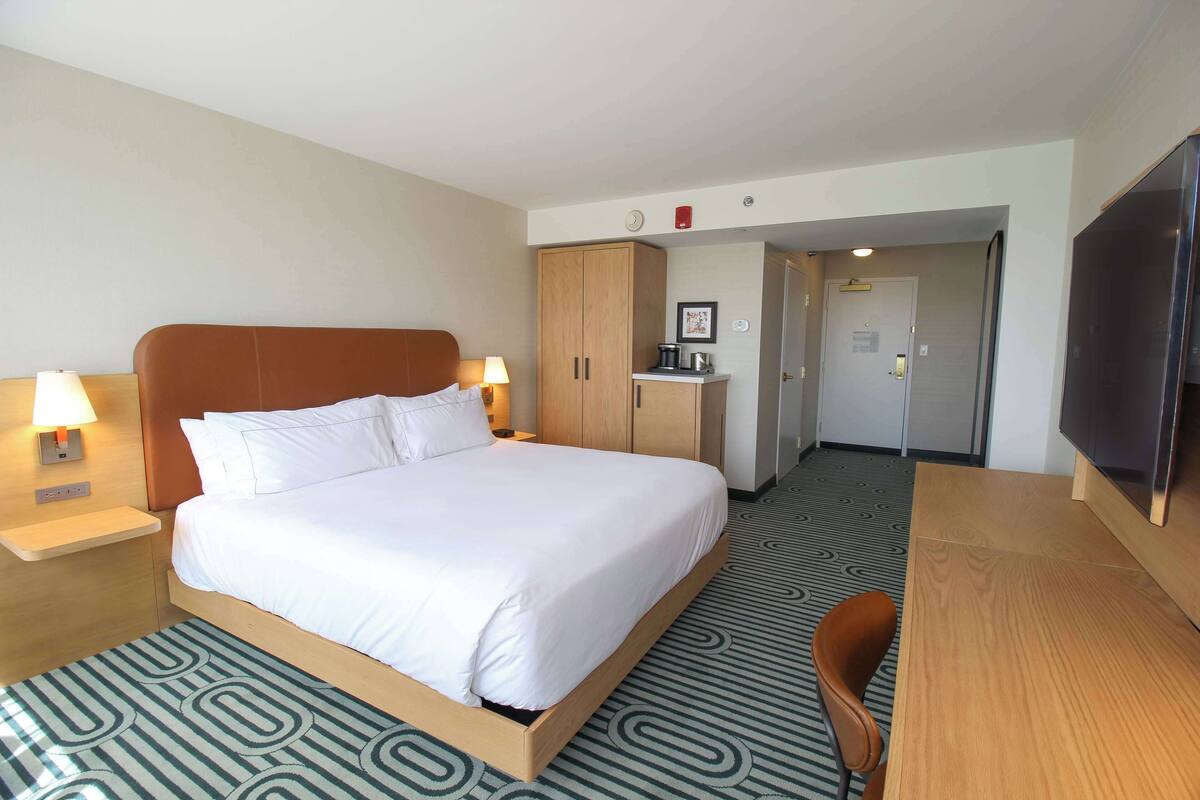 Une chambre d’hôtel avec un grand lit, des tables de chevet avec des lampes, un bureau en bois, une chaise, une télévision et un coin cuisine.