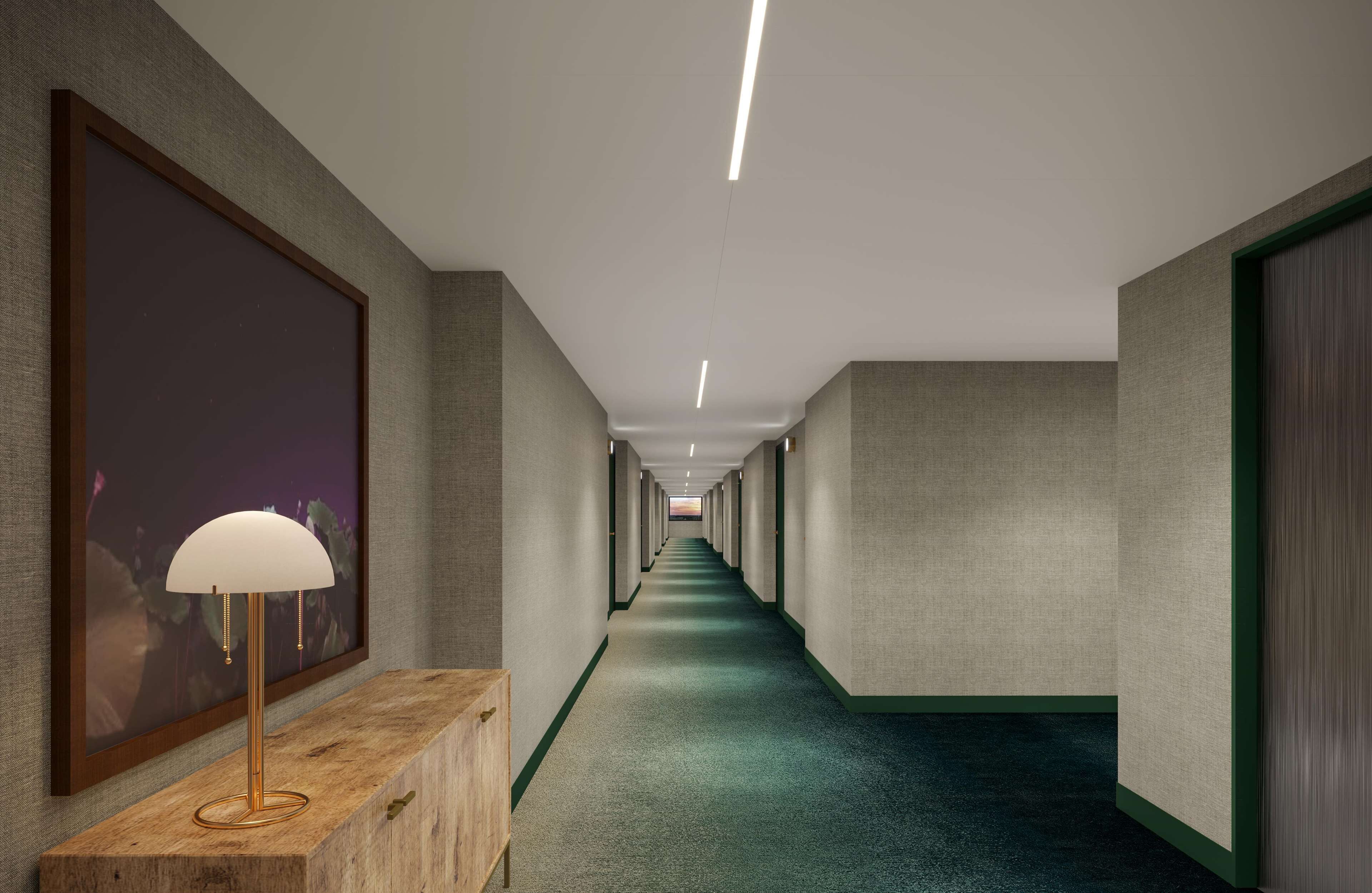 A long, narrow hallway with a green carpet, a wooden bench with a lamp, and framed artwork on the walls.