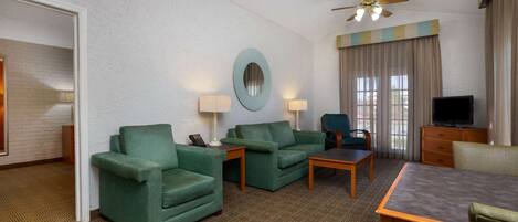 A hotel room with a green sofa set, a wooden coffee table, a TV, and a ceiling fan.