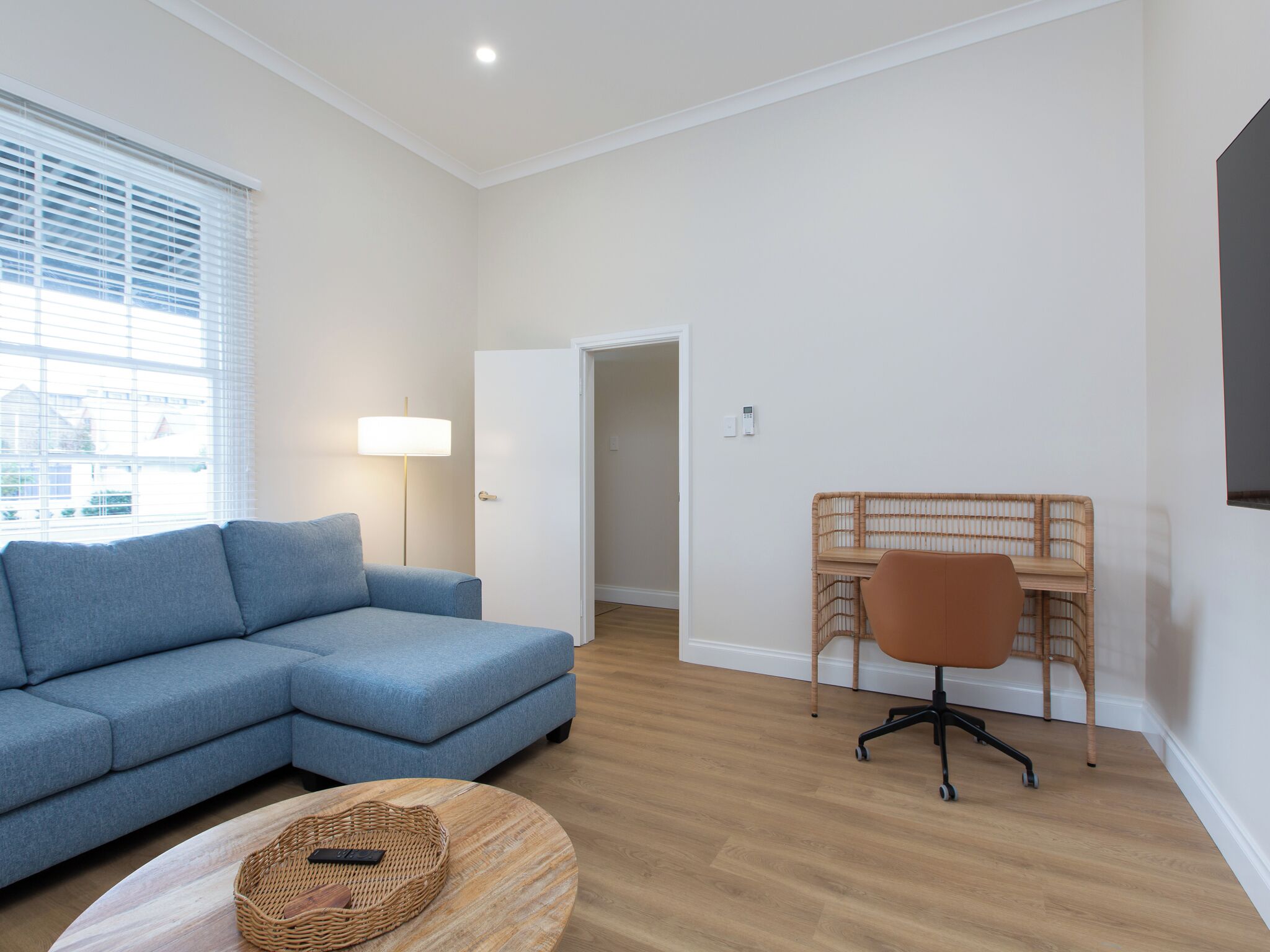 Una sala moderna con un sofá seccional, una mesa redonda de centro, una mesita de mimbre, un escritorio con silla y un gran ventanal con persianas.