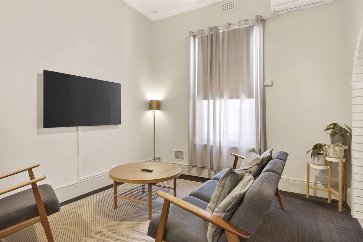 A modern living room with a flat-screen TV mounted on the wall, a round wooden coffee table, a grey sofa with wooden legs, and a small side table with a plant.