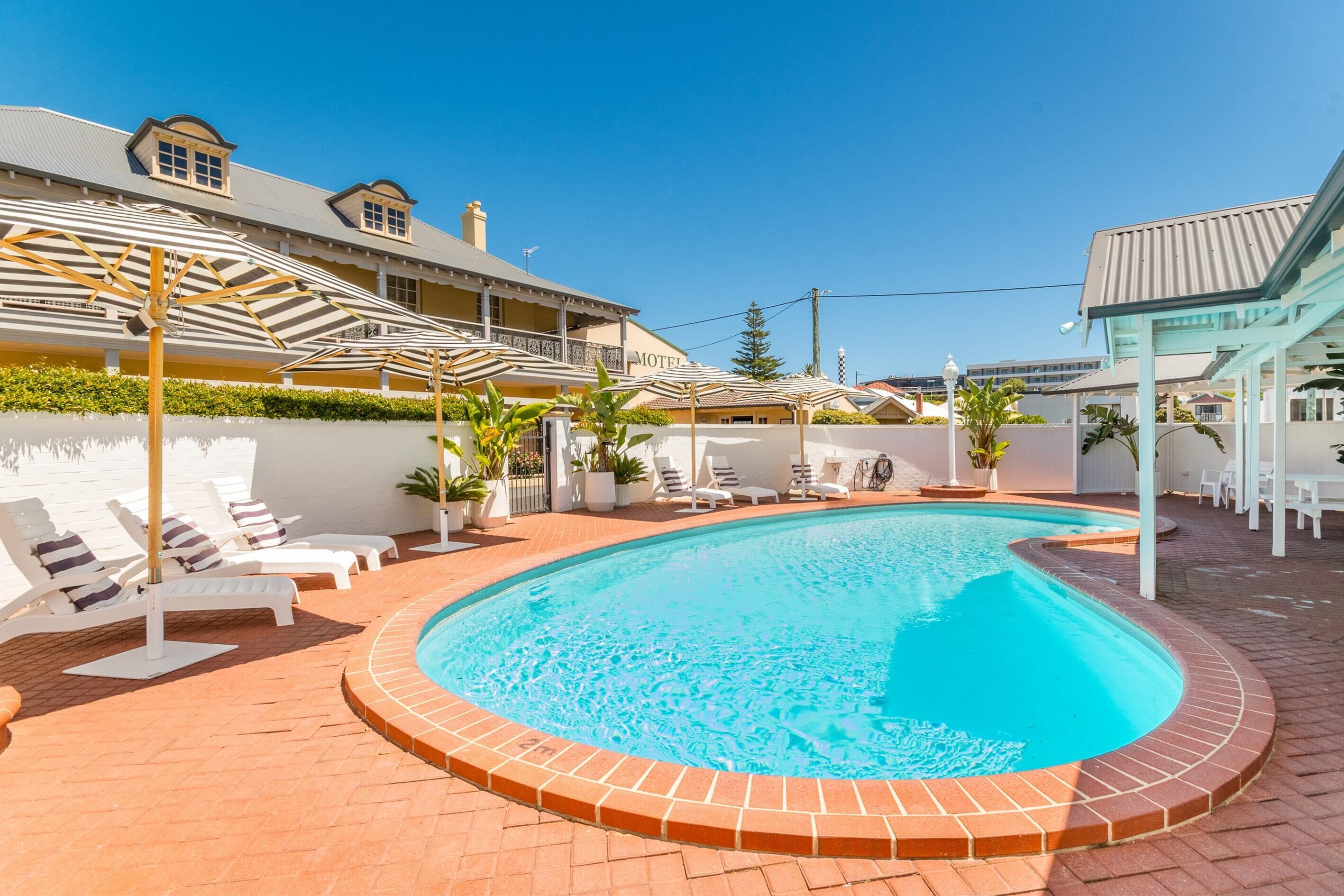 A swimming pool with sun loungers and umbrellas, surrounded by a brick patio and a building with a sign that reads "Motel".