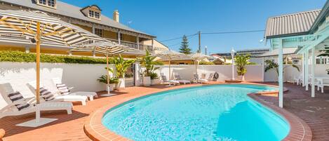 A swimming pool with sun loungers and umbrellas, surrounded by a brick patio and a building with a sign that reads "Motel".