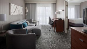 Habitación de hotel con sofá seccional gris, escritorio con silla, cama, televisor y ventana grande con cortinas translúcidas.