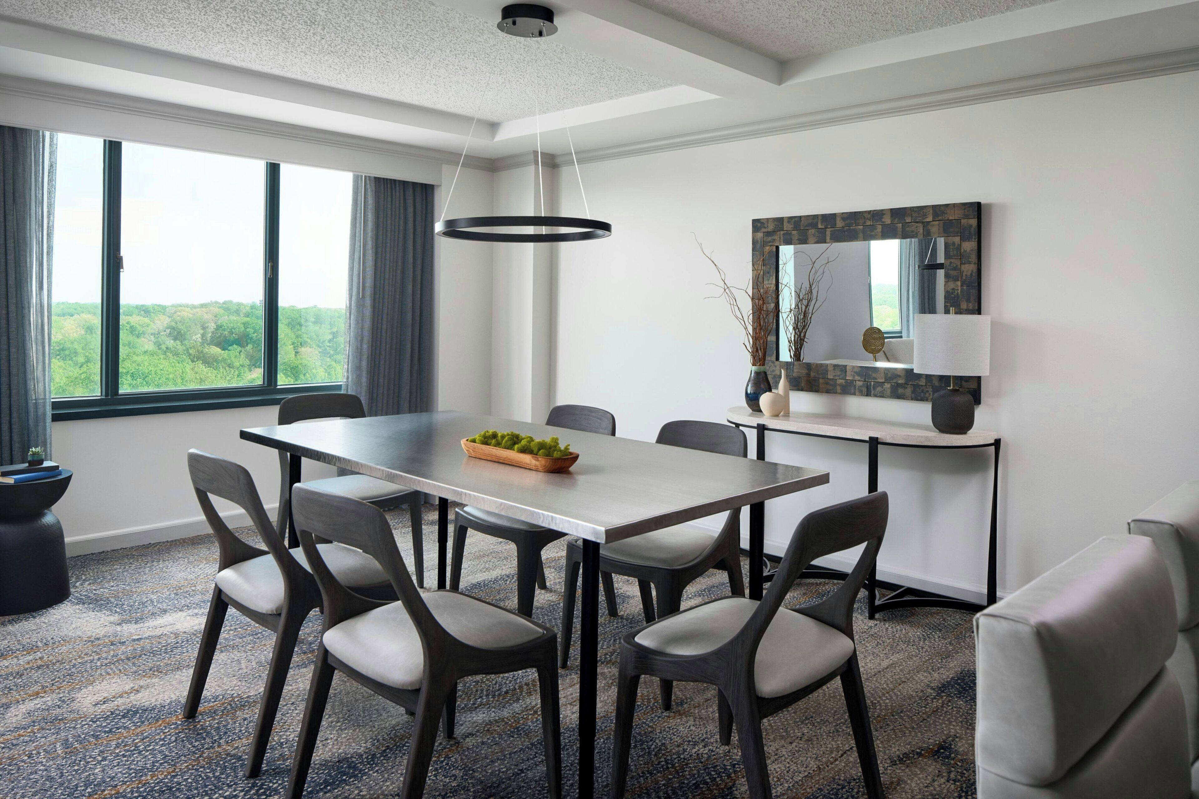 A modern dining room with a large table, six chairs, a hanging light fixture, a mirror, and a view of greenery outside.