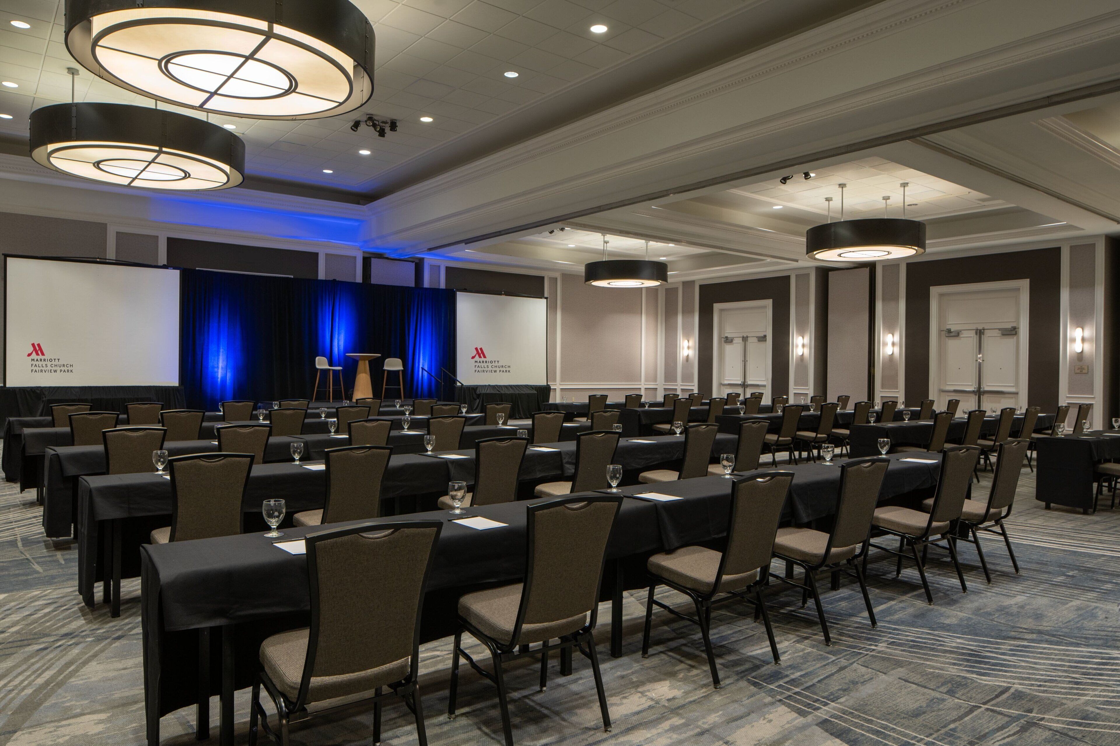 A conference room with rows of tables and chairs, a stage with a blue curtain, and a projector screen.
