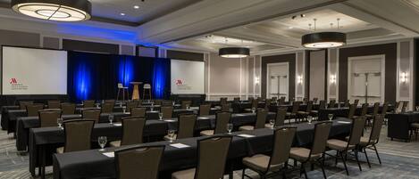 A conference room with rows of tables and chairs, a stage with a blue curtain, and a projector screen.