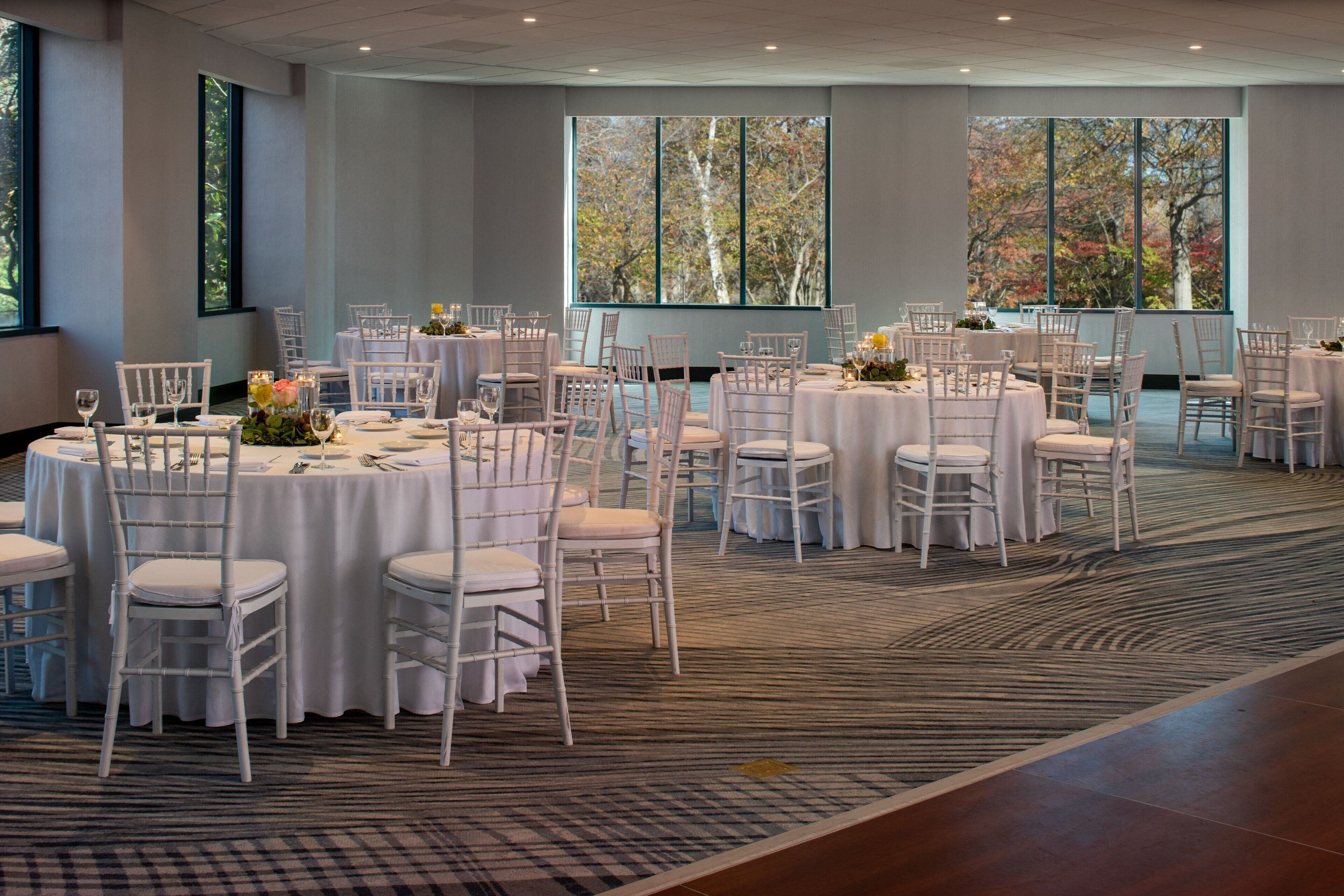 A banquet hall with round tables, white chairs, and large windows with autumn foliage views.