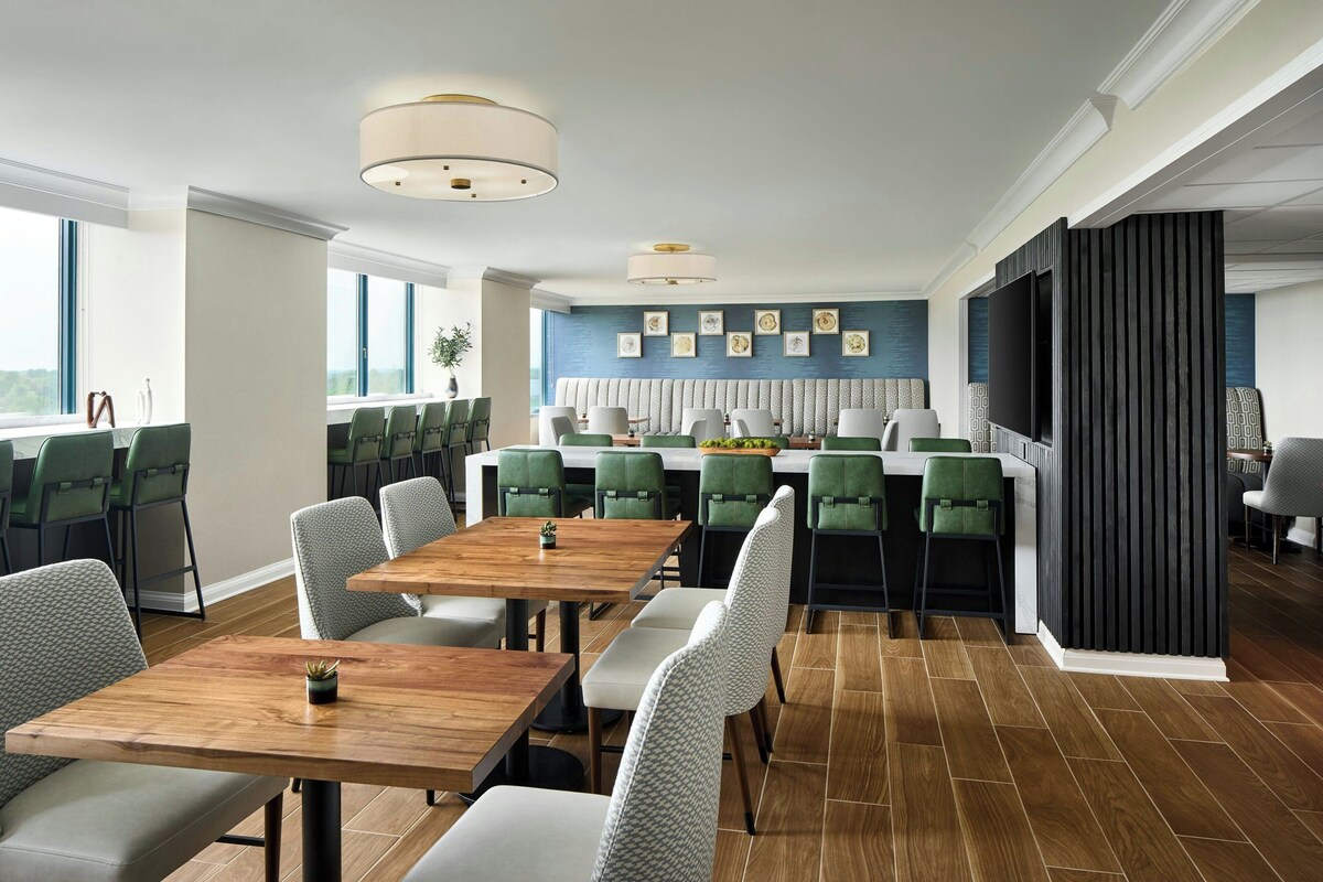 A modern dining area with wooden tables and green upholstered chairs, a kitchen with a large TV, and a view of the outdoors.