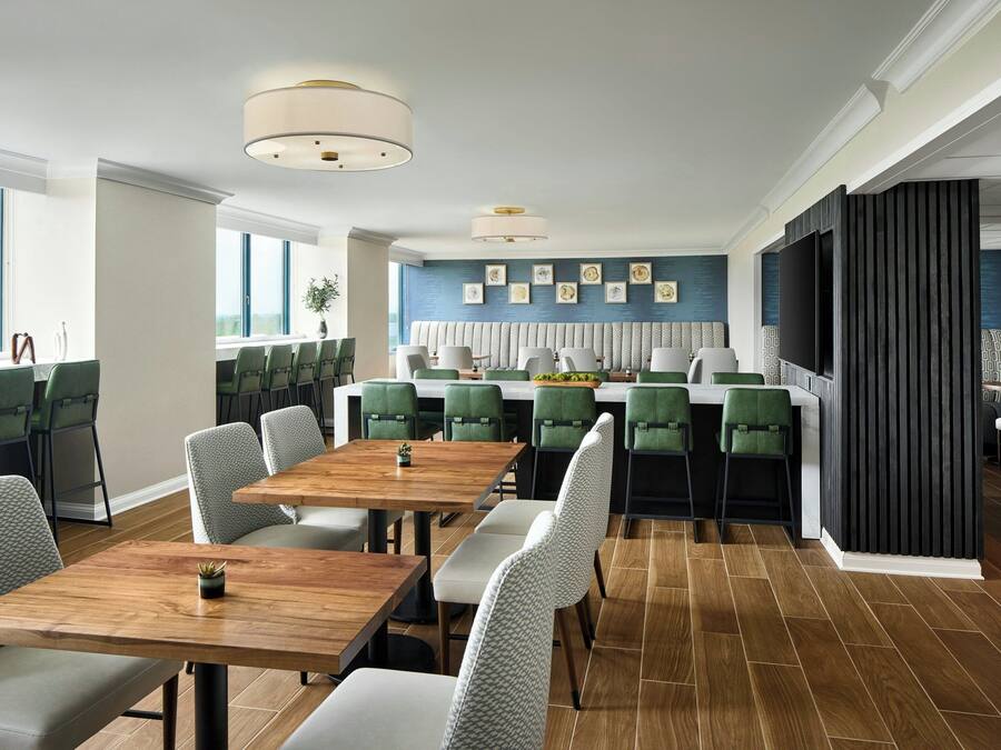 A modern dining area with wooden tables and green upholstered chairs, a kitchen with a large TV, and a view of the outdoors.