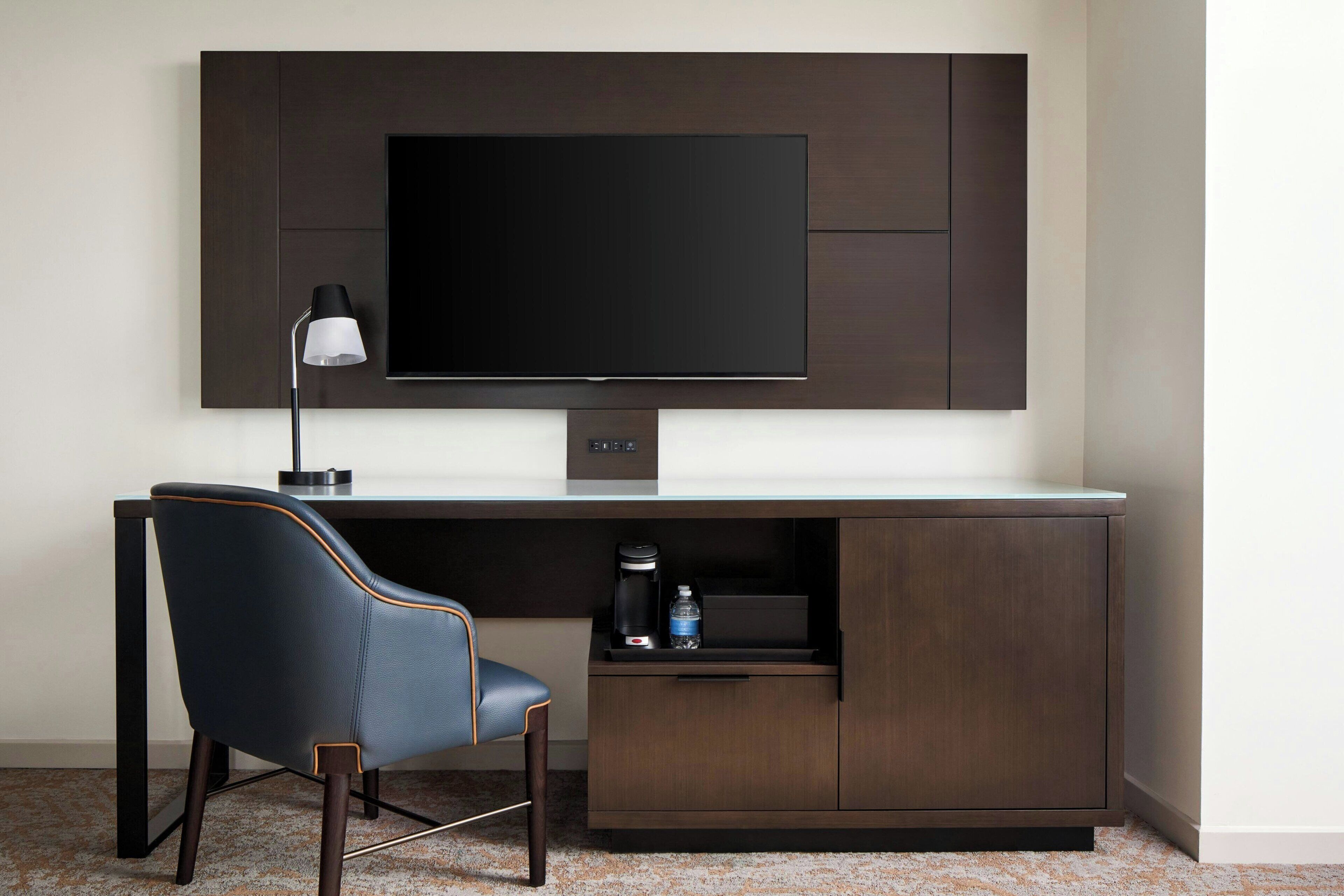 A modern room with a flat-screen TV mounted on a dark wooden wall, a desk with a chair, a coffee maker, and a water bottle on the desk.