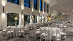Un salón de banquetes con mesas redondas y sillas blancas, ventanas amplias y jarrones decorativos.