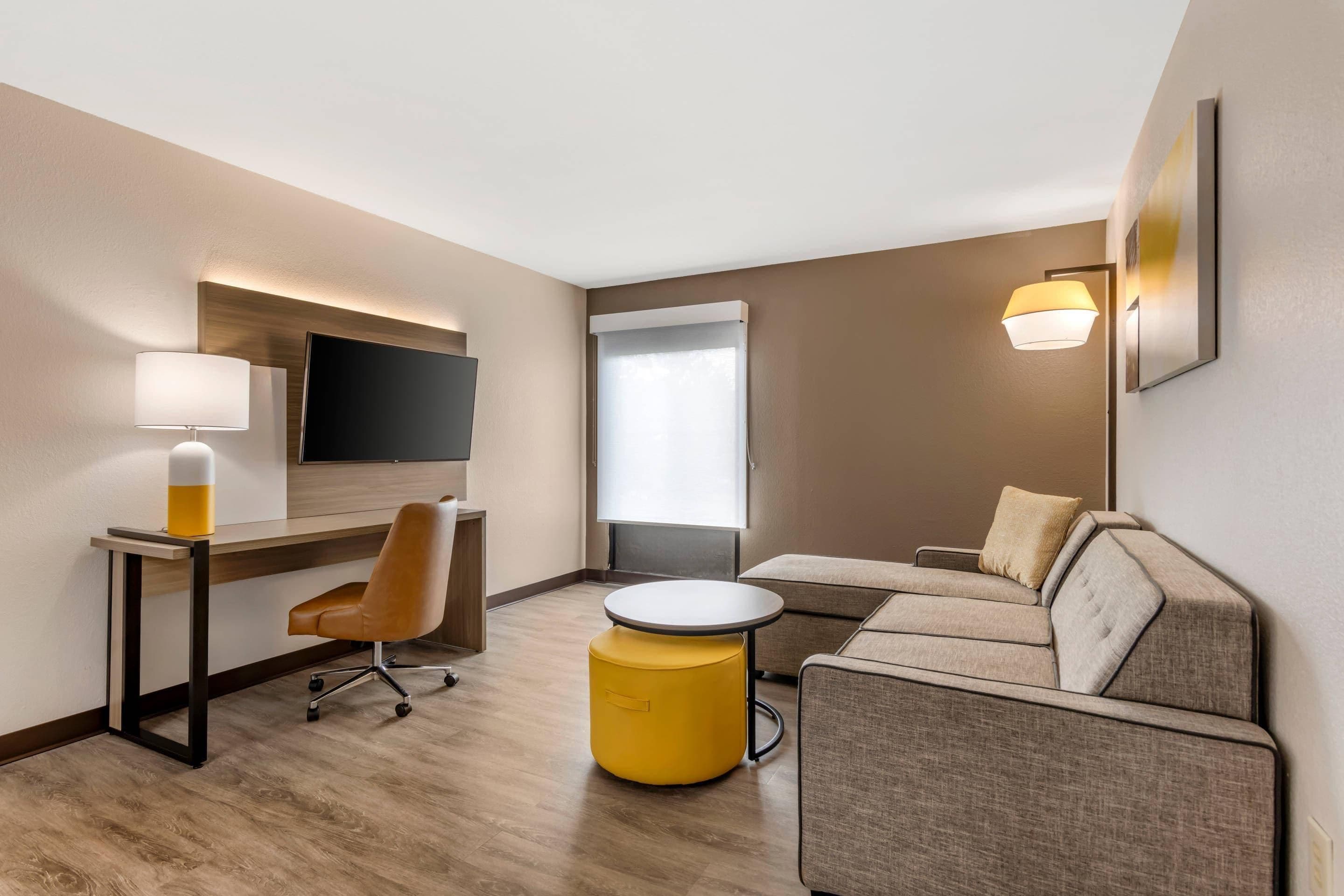 Una habitación de hotel moderna con una televisión de pantalla plana montada en la pared, un escritorio con una silla, un reposapiés amarillo redondo, un sofá y un ventanal grande que proporciona luz natural.