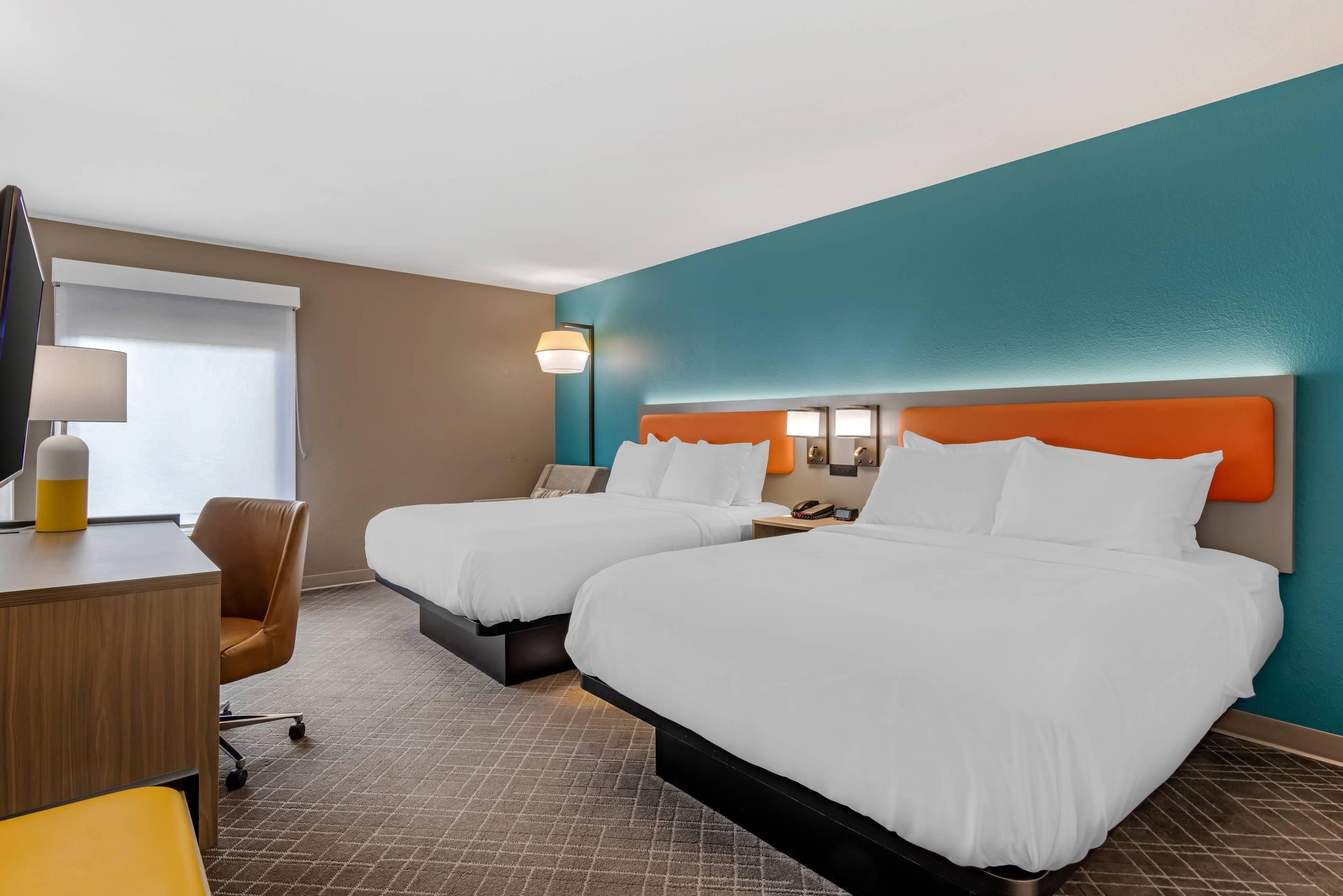 A hotel room with two beds, a desk, a chair, a lamp, and a window with blinds.