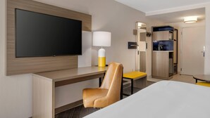 A hotel room with a large flat-screen TV mounted on the wall, a desk with a chair, a bedside table, a lamp, and a wardrobe.