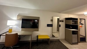 A hotel room with a desk, chair, yellow bench, mirror, wardrobe, and a TV.