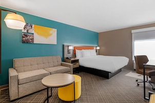 Una habitación de hotel con cama, sofá, una mesita redonda, lámpara y cuadros en la pared.