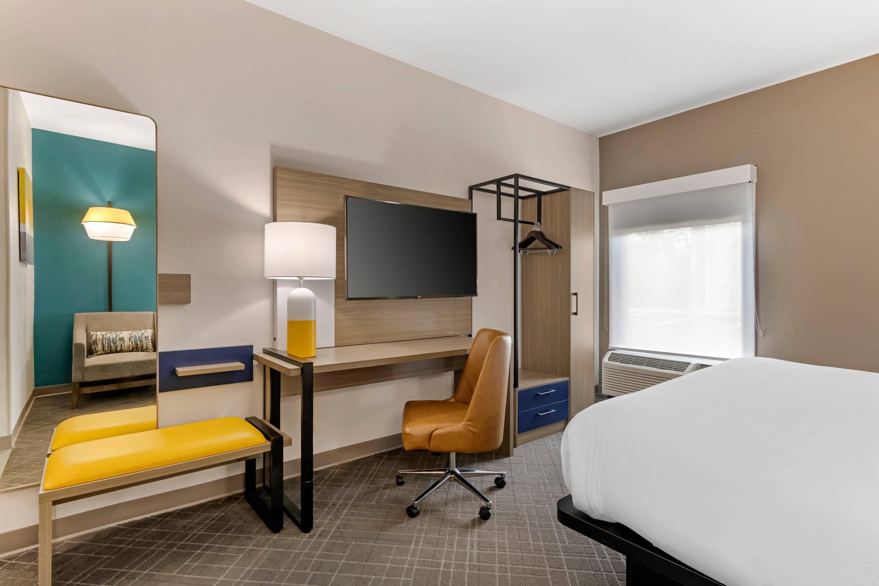 Ein Hotelzimmer mit einem Bett, einem Schreibtisch mit Stuhl, einer Bank, einem Fernseher, einem Spiegel und einem Fenster mit Vorhängen.