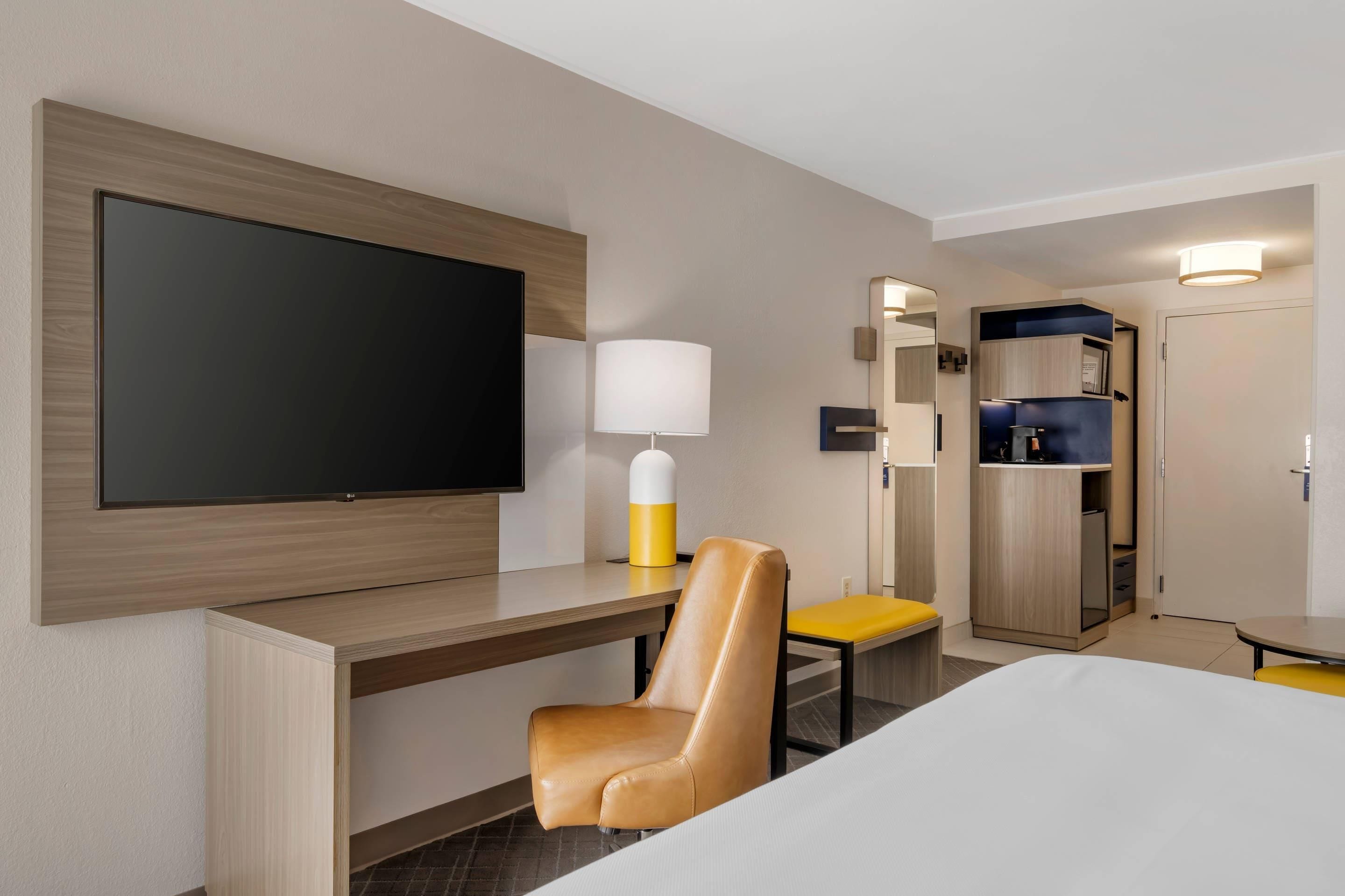 A hotel room with a large flat-screen TV mounted on the wall, a desk with a chair, a bedside table, a lamp, and a wardrobe.