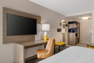 Habitación de hotel con una televisión de pantalla plana grande montada en la pared, un escritorio con silla, una mesita de noche, una lámpara y un armario.