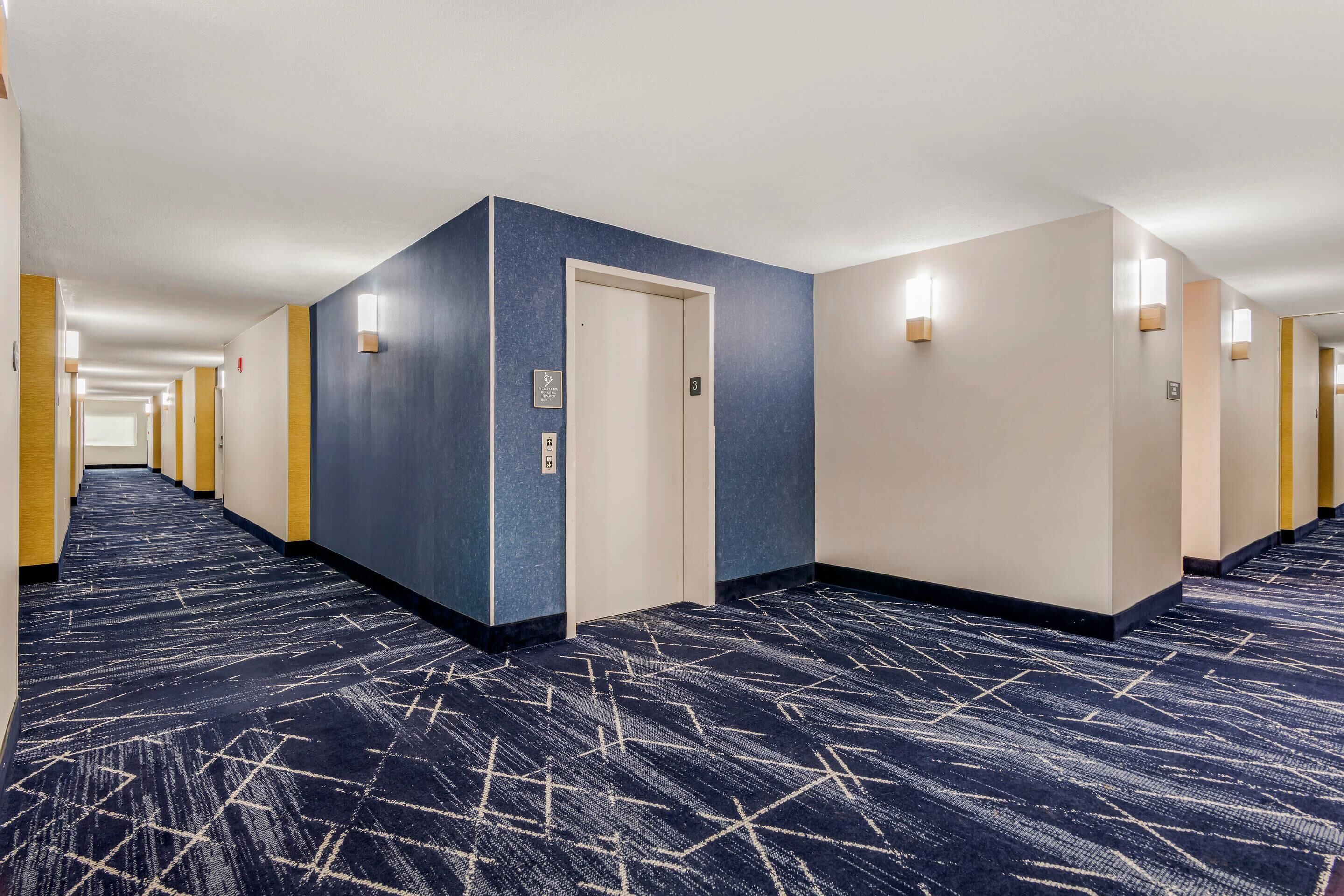 Un pasillo de hotel con alfombra azul, paredes de colores claros y una puerta con señal de ascensor.