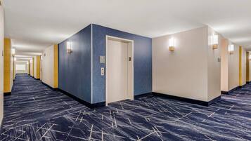 Un pasillo de hotel con alfombra azul, paredes de colores claros y una puerta con señal de ascensor.