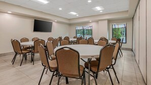 A conference room with multiple tables and chairs, a flat-screen TV, and large windows with views of greenery.