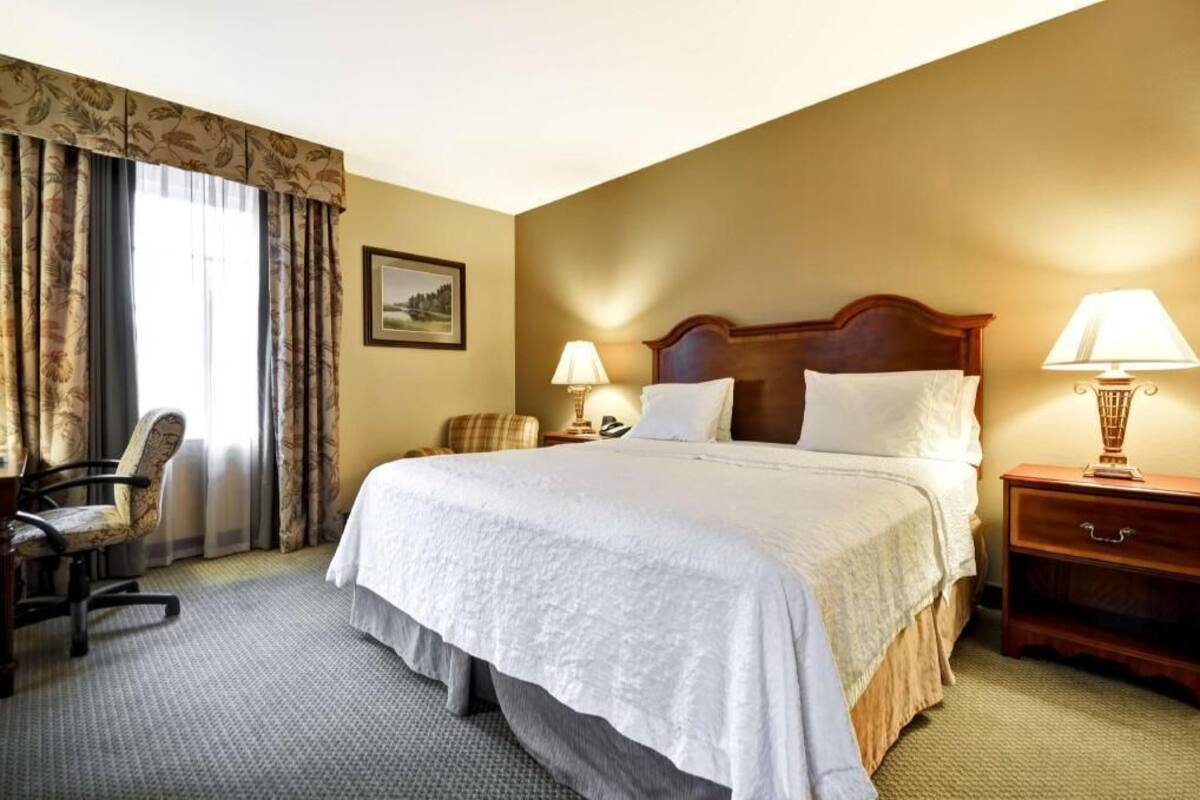 A hotel room with a large bed, two bedside lamps, a chair, a desk, and a picture on the wall.