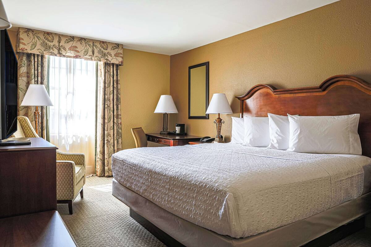 A hotel room with a large bed, two bedside lamps, a desk with a lamp, a chair, and a large window with curtains.